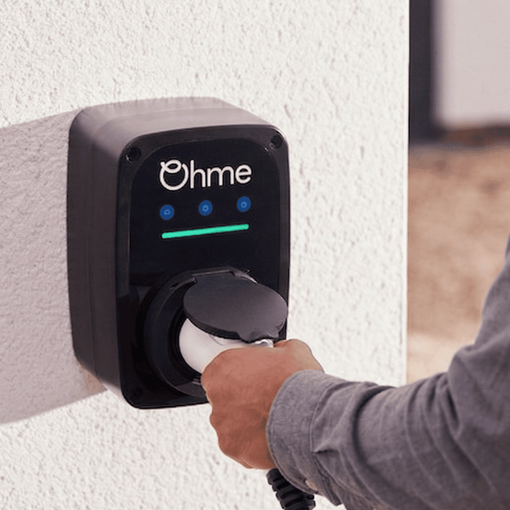 Hand inserting the Type 2 connector into an Ohme smart EV charger mounted on a UK home exterior wall, an OZEV-approved chargepoint eligible for the EV Chargepoint Grant