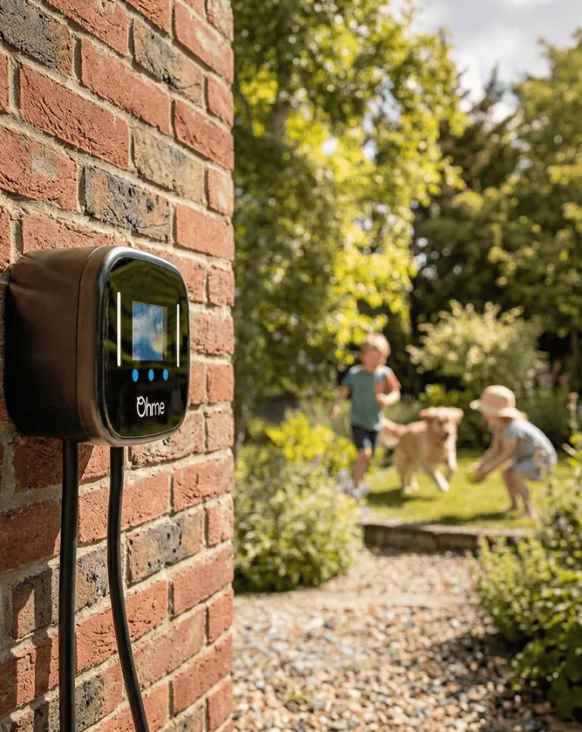 Ohme home EV charger on a brick wall, with a family and dog in the garden behind