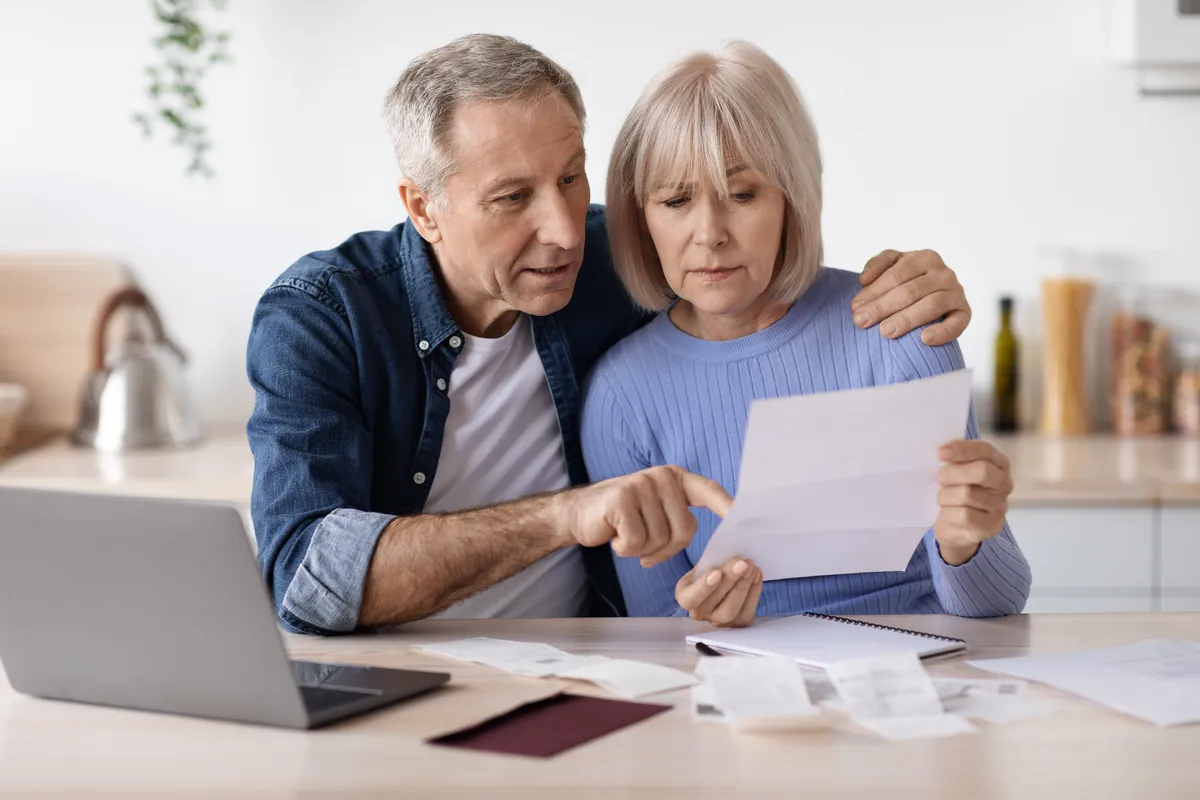 Older couple at a kitchen table reading a letter together beside a laptop