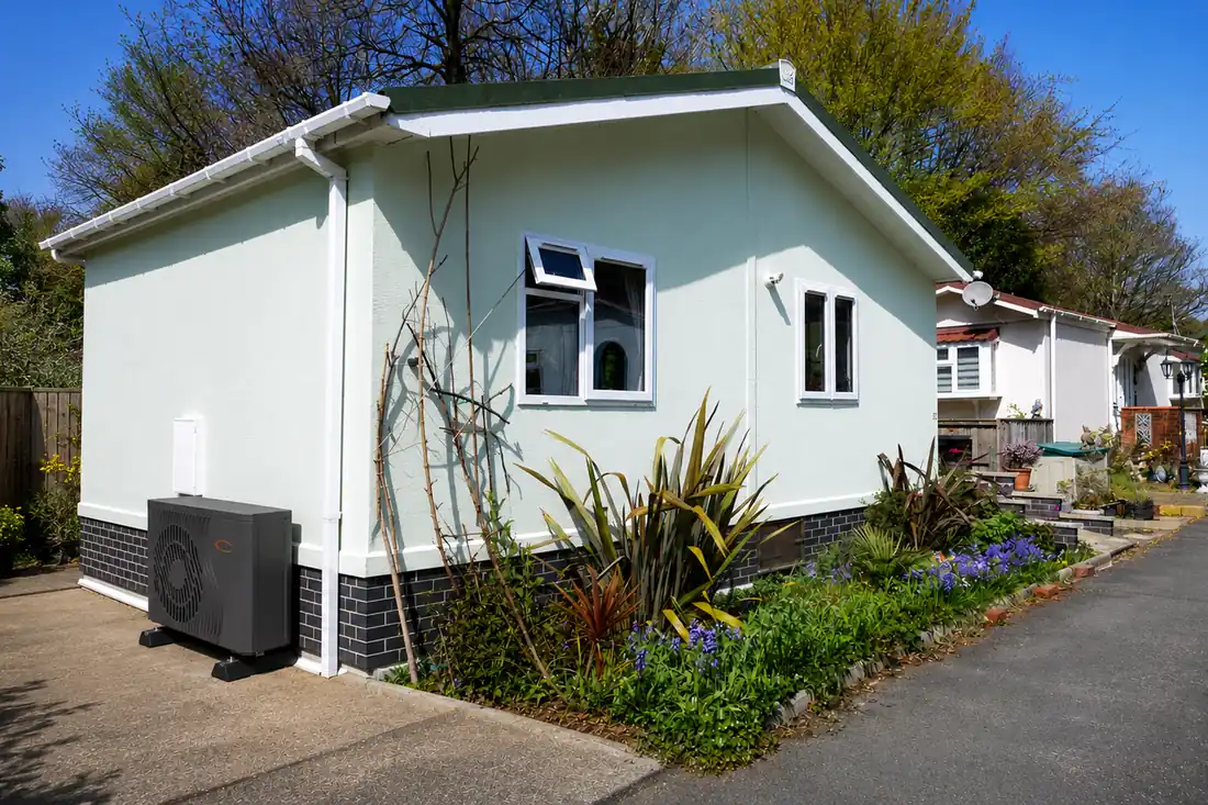 Air source heat pump on a concrete pad beside a residential park home, with neighbouring park homes along the street