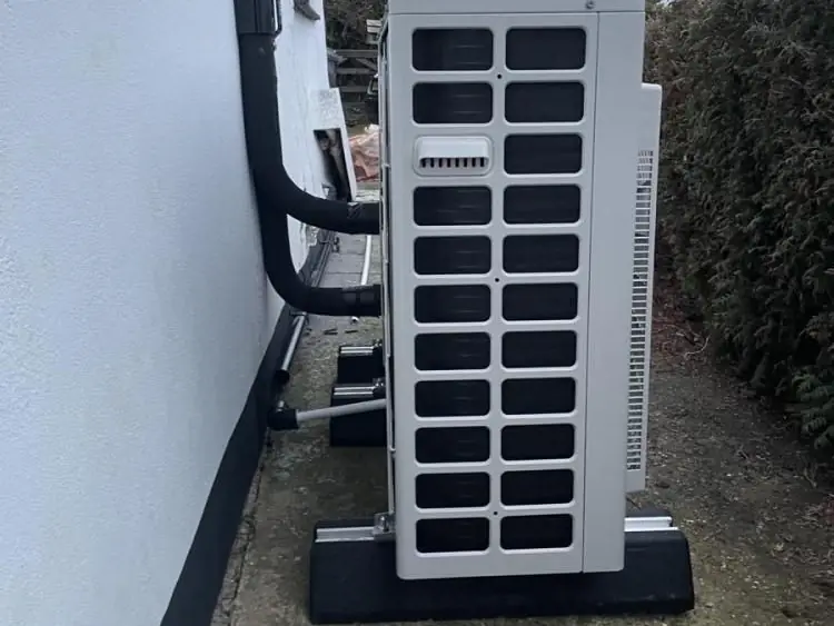 Side view of an outdoor heat pump unit on a plinth, with insulated pipework running into the house