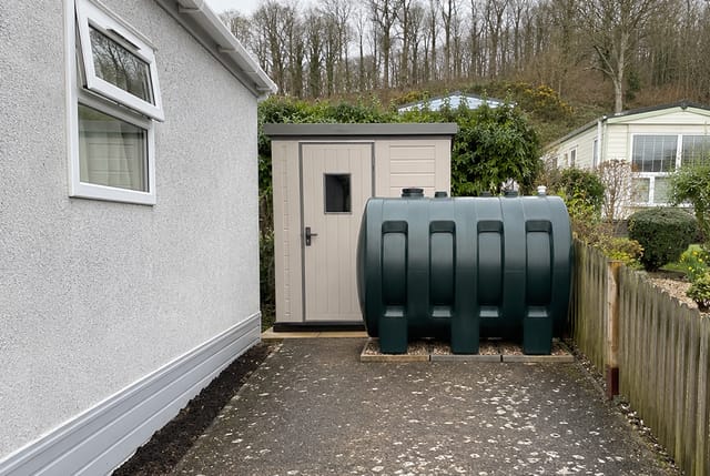 Green outdoor oil heating tank on a park home plot, the delivered fuel a heat pump replaces