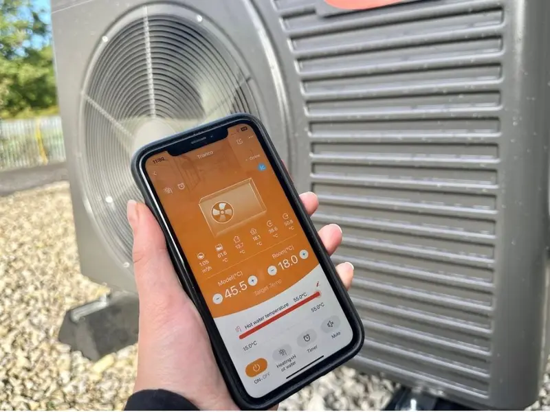 Hand holding a phone running the Trianco app in front of an Activair air source heat pump