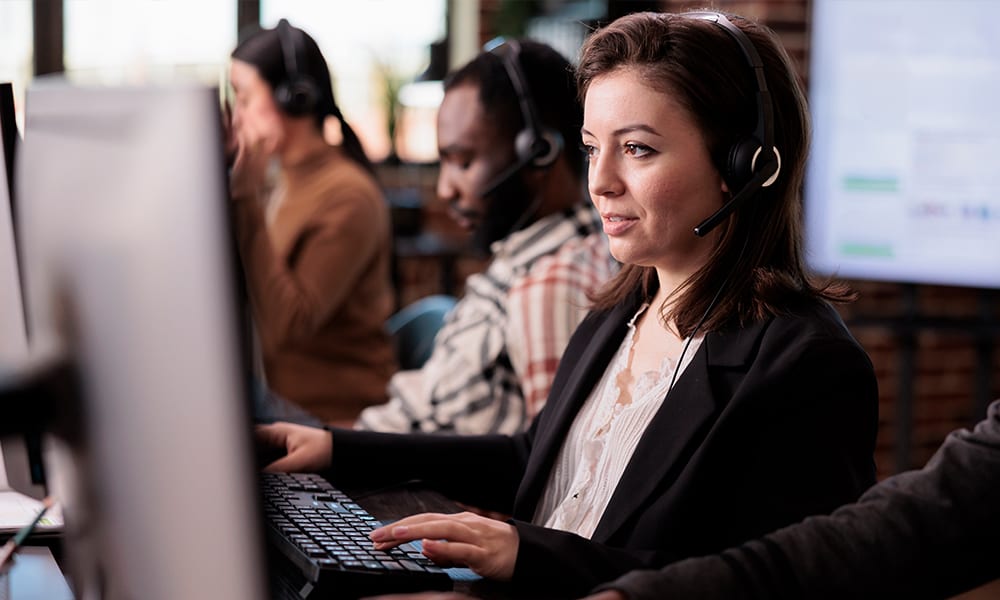 Focused customer service advisor with a headset typing at her desk in a modern, bright office with colleagues in the background